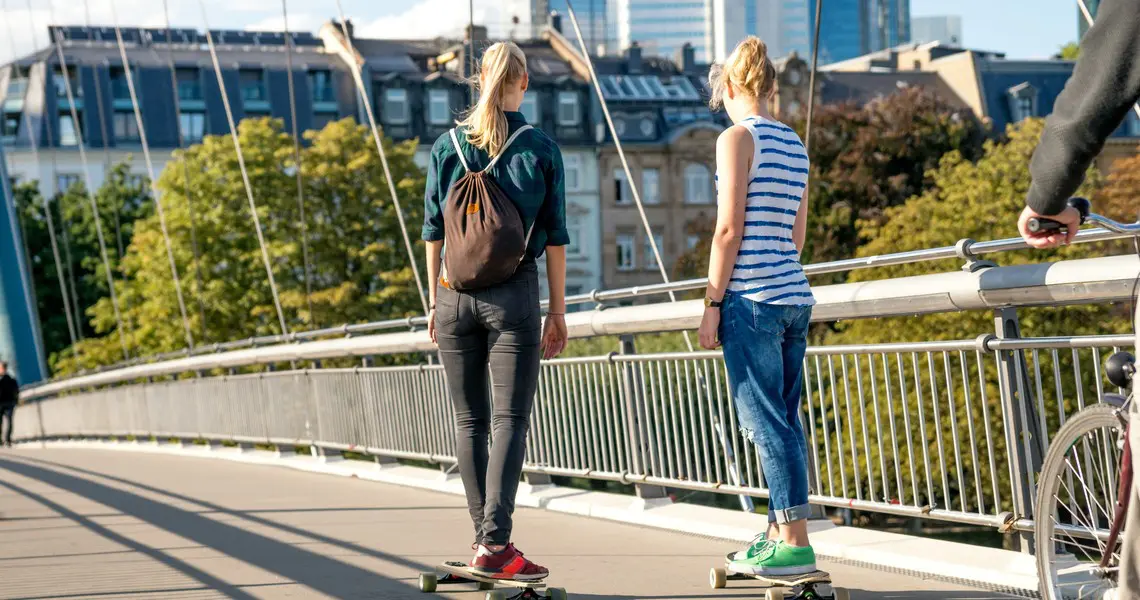 News 7 Zwei junge Frauen fahren auf Longboards über einen Steg vor der Frankfurter Skyline